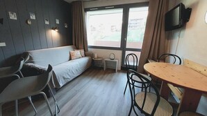 Ein Zimmer mit einer Couch, einem kleinen Tisch und Stühlen. Ein Fernseher ist an der Wand montiert und es gibt ein Fenster mit Vorhängen.