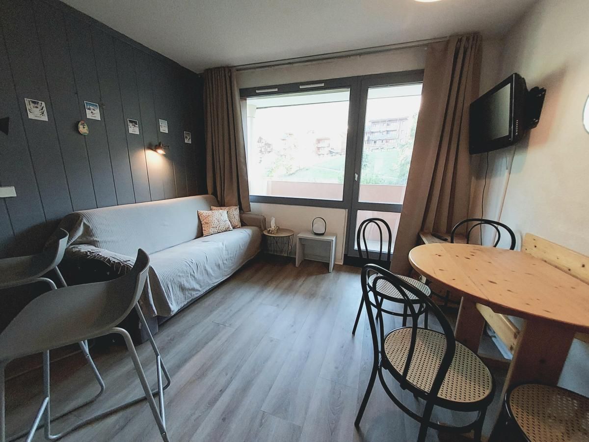 A room with a sofa, a small table, and chairs. There is a TV mounted on the wall and a window with curtains.