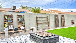 A backyard with a fire pit, potted plants, and a wooden shelf with hanging lights.