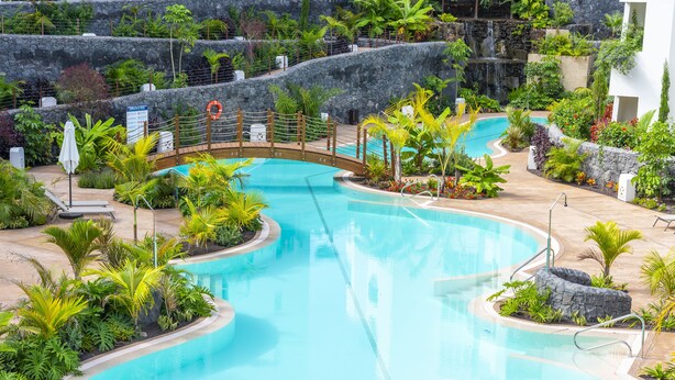 A large, clear swimming pool surrounded by tropical plants and a stone wall. - Luxury apartment: air conditioning, swimming pools, heating, gym, fibre optics, garage (Palm-Mar)