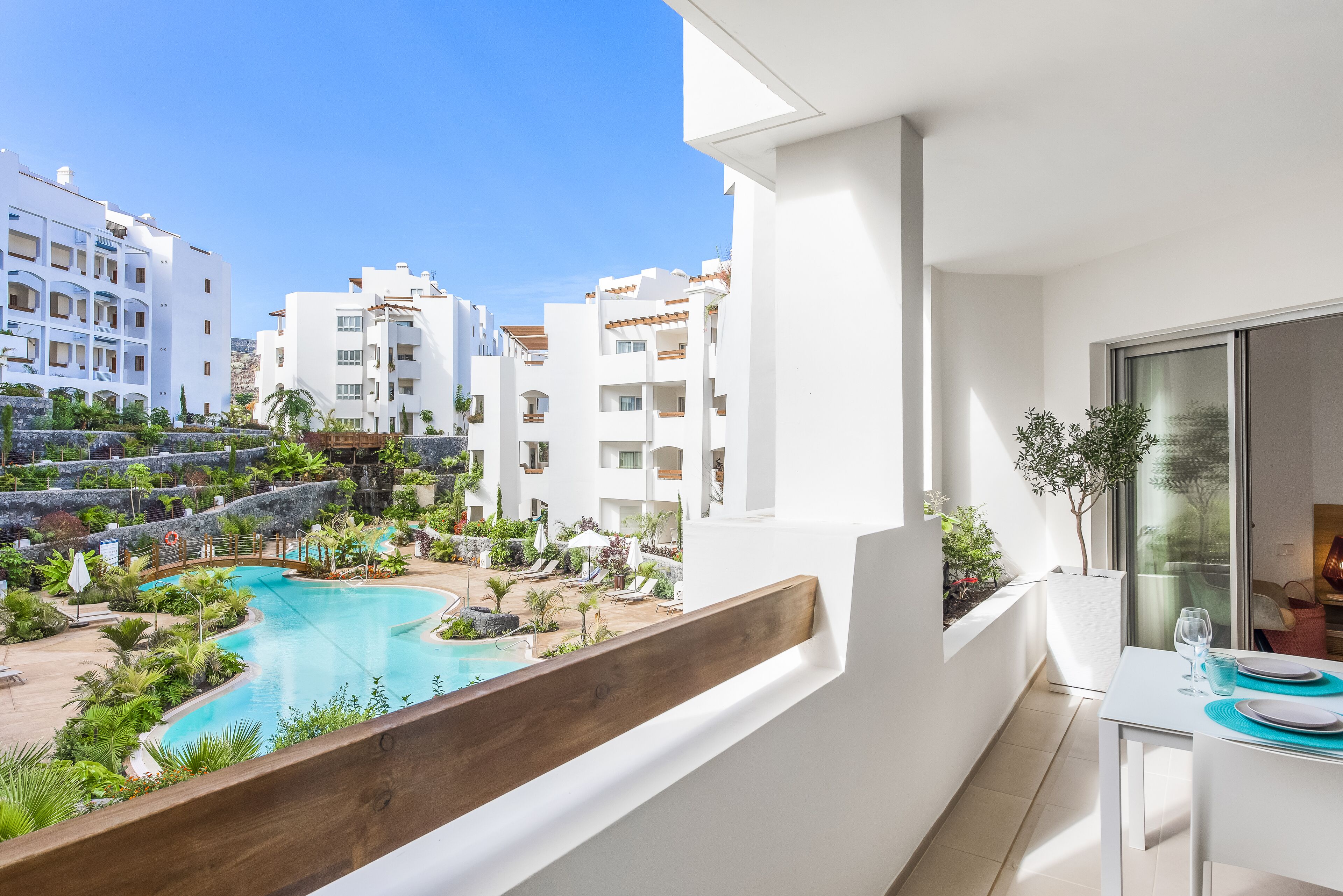 A balcony with a pool, white buildings, and a dining area with a table and chairs.