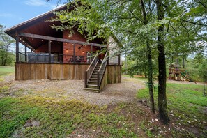 Une cabane en bois avec une terrasse couverte, un escalier et une aire de jeux en arrière-plan.