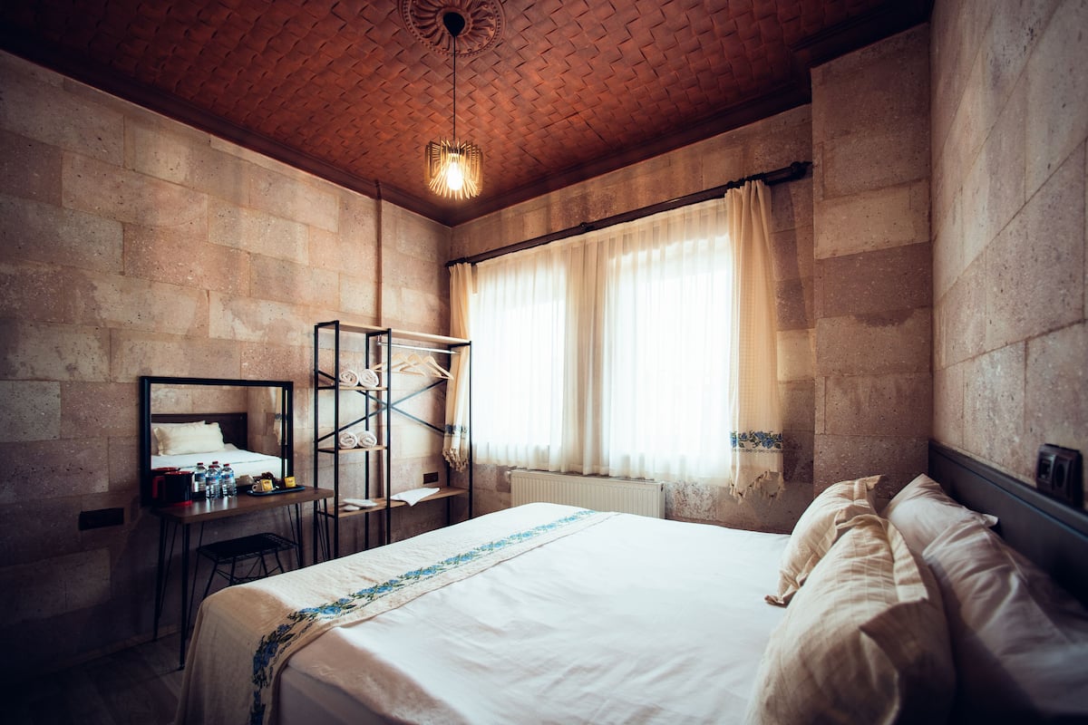 A hotel room with a brick ceiling, a bed with white linens, a desk with a mirror, and a window with curtains.
