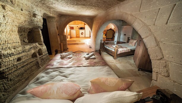 A stone-walled room with a bed, a wooden chest, and a small table. There are two arched doorways leading to another room with a bed and a mirror. - Azalea Terrace Cave Hotel (Nevsehir)