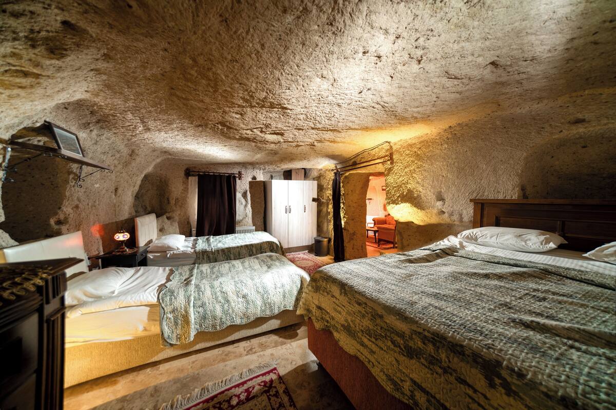 A rustic, cave-like bedroom with two beds, a wooden wardrobe, and a small table with a lamp.