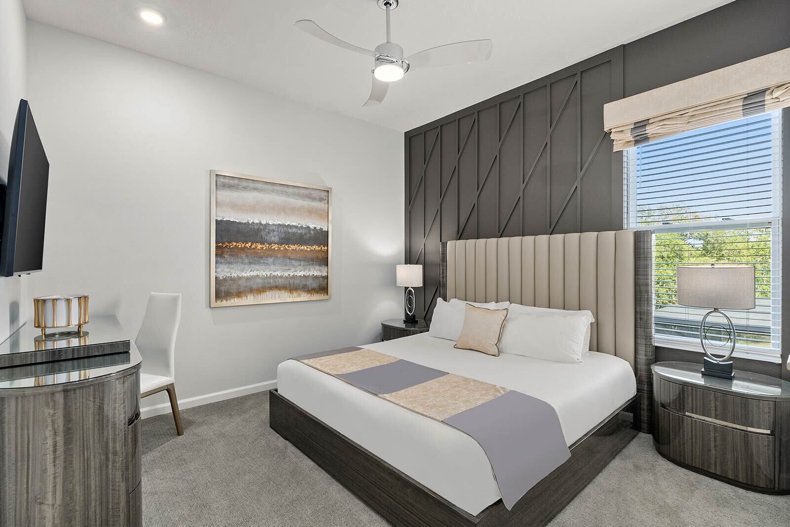 A modern hotel room with a large bed, a ceiling fan, a painting, a desk with a lamp, and a window with blinds.