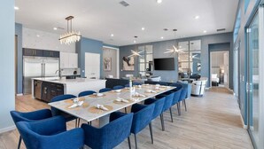 Une cuisine moderne avec des meubles sombres et un grand îlot central, un coin repas avec des chaises bleues et une table dressée pour six personnes, et un salon avec un téléviseur et des fauteuils.