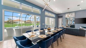 A modern dining area with a long table, blue chairs, and large windows offering a view of a pool and greenery. - Action-Packed Home w Sports Simulator, Bowling, Gamer BR & Theater - Near Parks (Davenport)