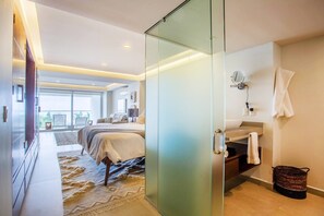A modern hotel room with a glass partition, a bed, a bathroom with a towel rack, and a view of the outdoors.