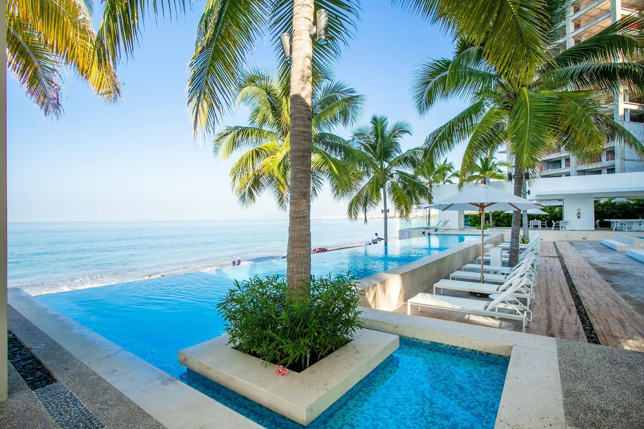 A pool area with a view of the ocean, surrounded by palm trees and lounge chairs.