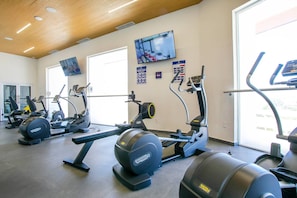 Et fitnessrum med forskellige træningsmaskiner, et tv monteret på væggen og et stort vindue, der giver naturligt lys.
