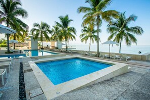 En swimmingpool med liggestole og parasoller, omgivet af palmer, med udsigt til havet.