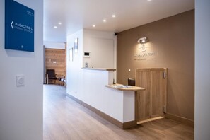 Un espace d’accueil avec un comptoir de réception en bois, une enseigne indiquant « Résidence Le Val Chavie » et un panneau bleu indiquant « DÉPÔT DE BAGAGES ».