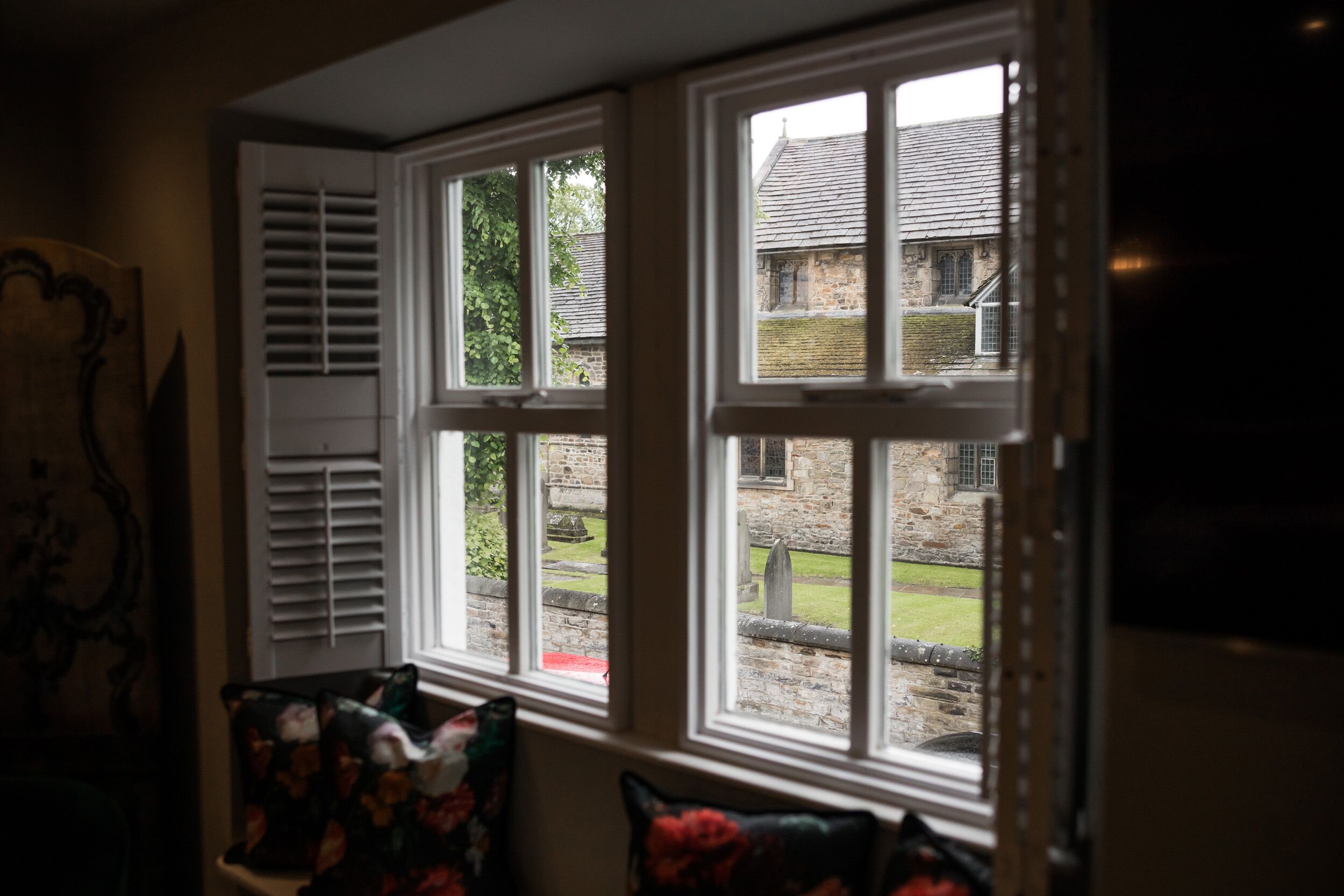 A room with a bay window overlooking a stone building and a grassy area.