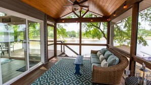 A covered outdoor area with a ceiling fan, a wicker sofa with cushions, a patterned rug, and a view of a body of water through large windows.