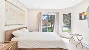 Un dormitorio con cama, mesita de noche, ventana con cortinas y vistas a árboles.