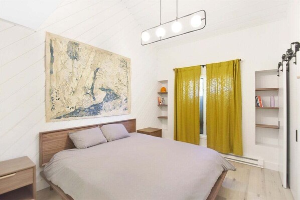 A bedroom with a large bed, a wooden headboard, a nightstand, a shelf with books, a window with yellow curtains, and a wall-mounted light fixture.