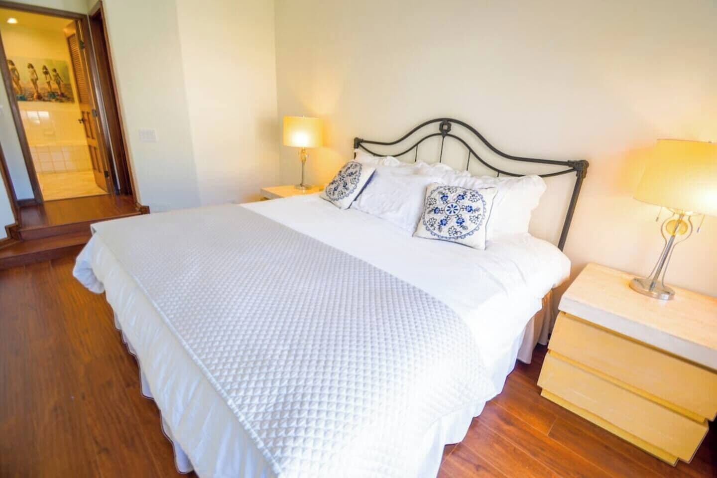 Una cama bien tendida con colcha blanca, dos almohadas decorativas, una lámpara de noche y suelo de madera.