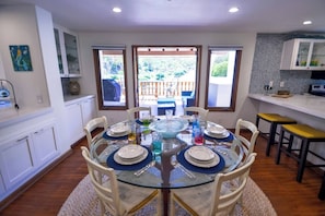 Comedor con mesa de vidrio, sillas blancas y una zona de cocina con encimera y armarios blancos.