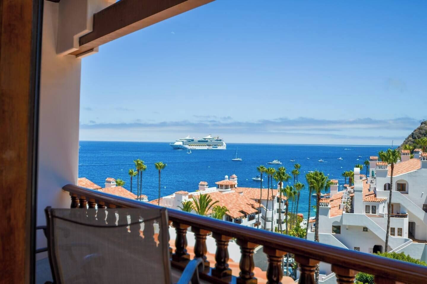 A balcony with a view of the sea, palm trees, and buildings with red-tiled roofs.