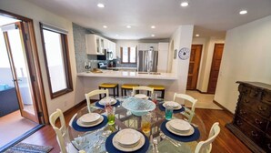 A dining area with a glass table set for six, surrounded by white chairs. A kitchen is visible in the background with modern appliances. - Oceanfront view, plus corner patio and golf cart (Avalon)
