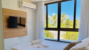 Habitación de hotel con cama, televisor montado en la pared y ventana con vista a palmeras y follaje.