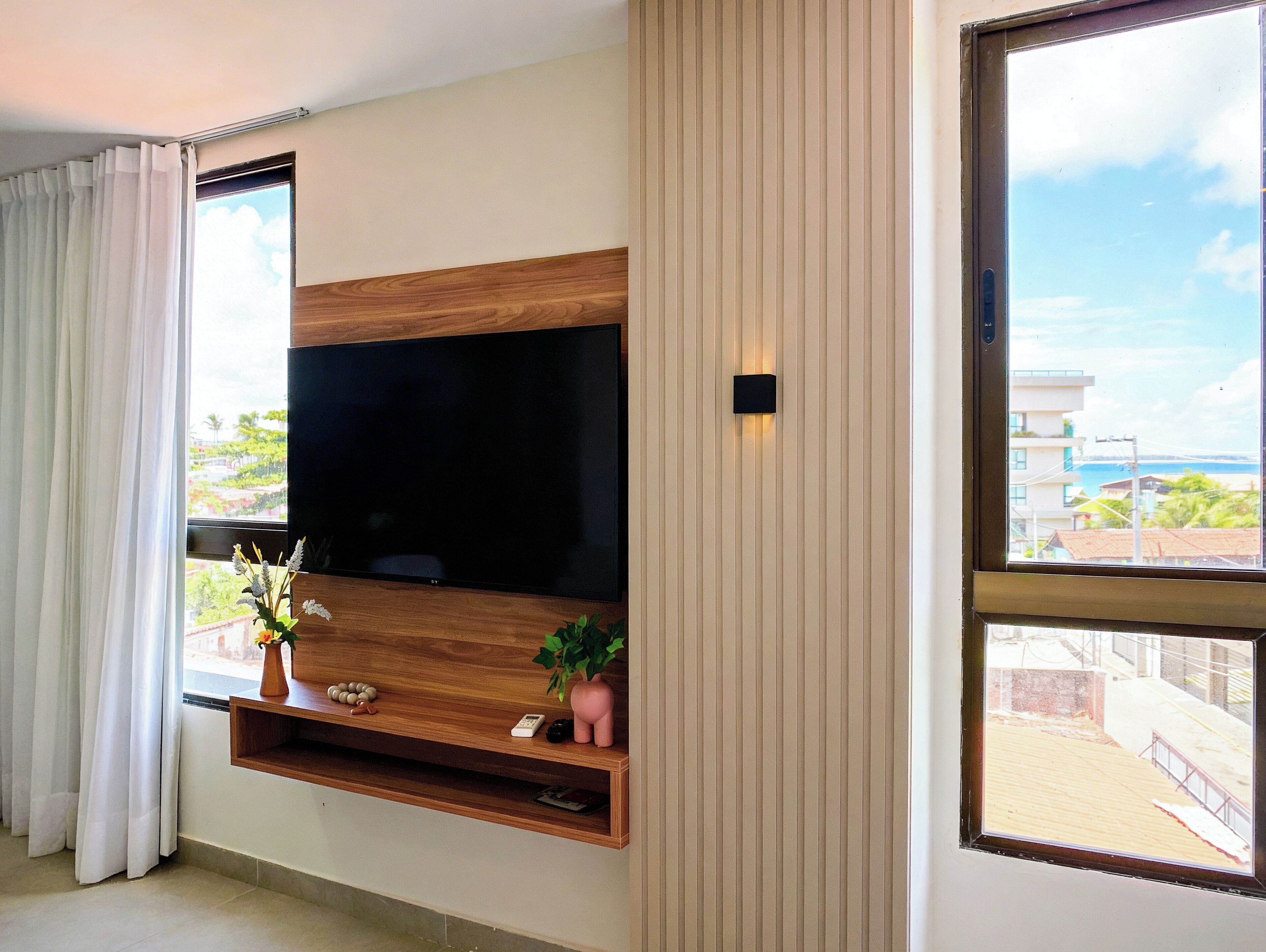 Ein modernes Wohnzimmer mit einem großen Flachbildfernseher an einer Holzvertäfelung, einem kleinen Regal mit Dekoration und Blick auf eine Küstenlandschaft durch ein großes Fenster.