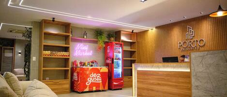 Een moderne hotellobby met een receptiebalie, een neonbord voor een minimarkt, en een Coca-Cola koelkast.