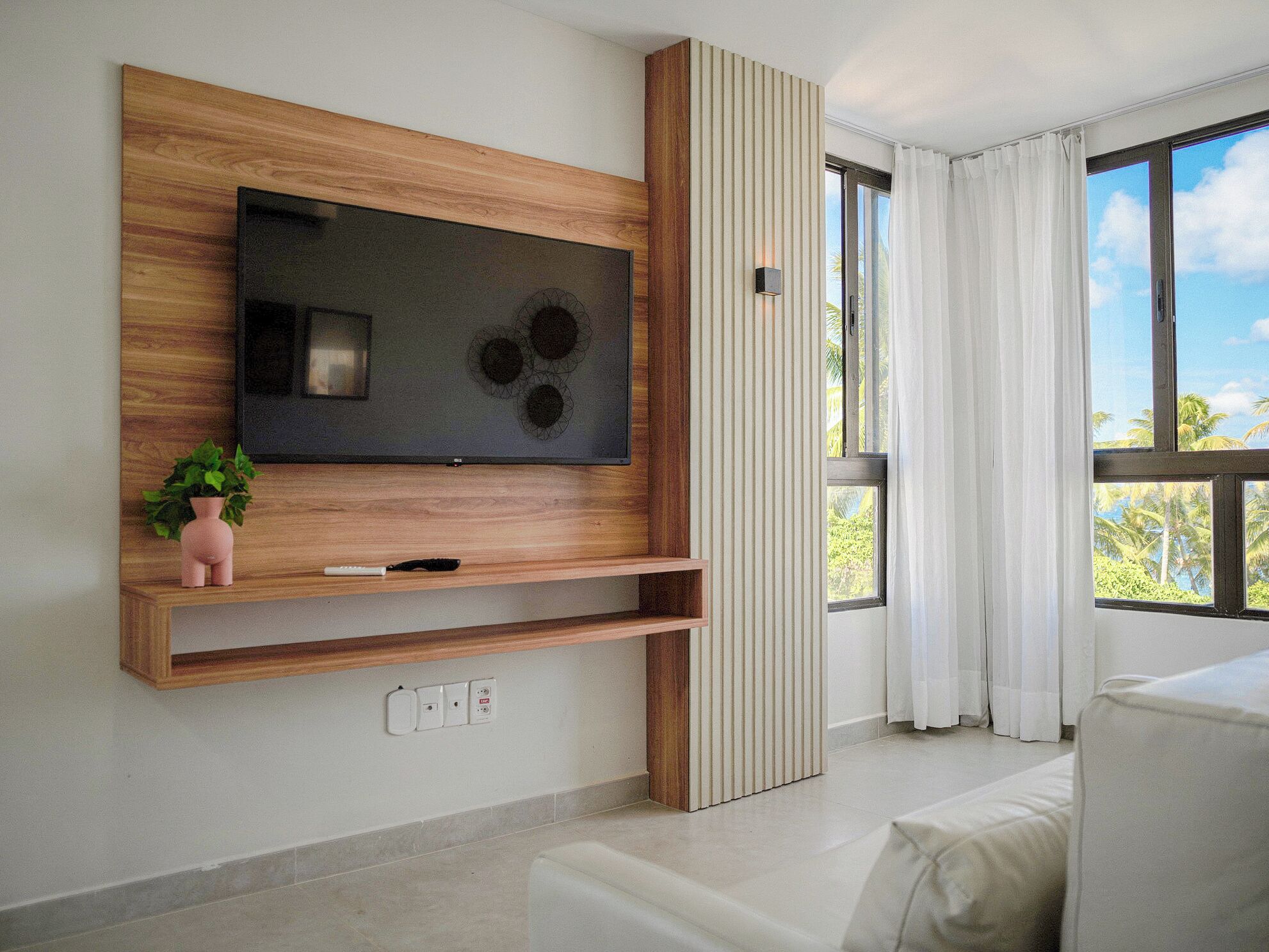 Una sala de estar moderna con un televisor de pantalla plana grande montado en una pared de madera, una pequeña planta rosa y un sofá blanco.