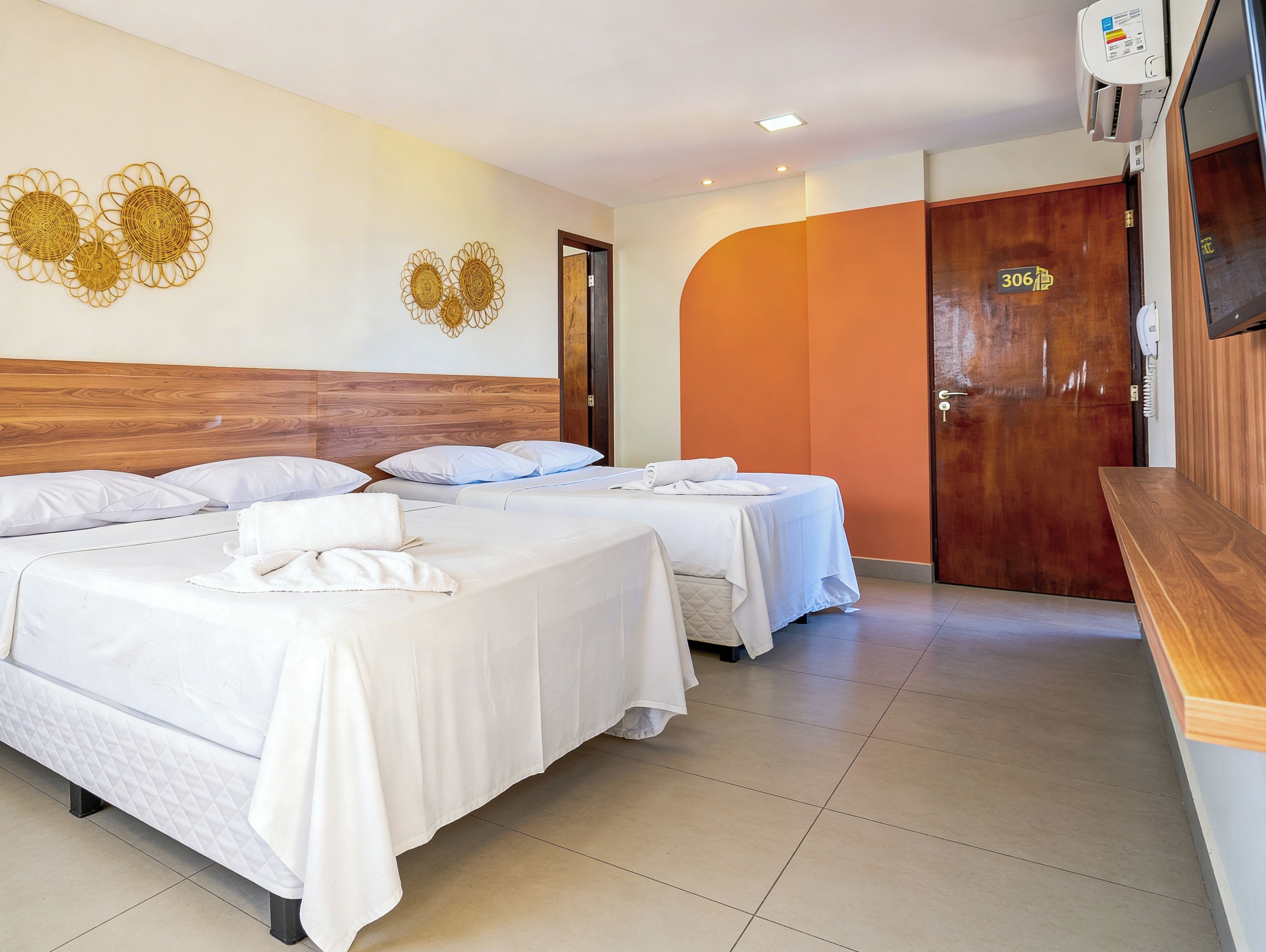 A hotel room with two beds, a wooden headboard, a wall with decorative wall art, a wooden door, and a reception area with a wooden counter.