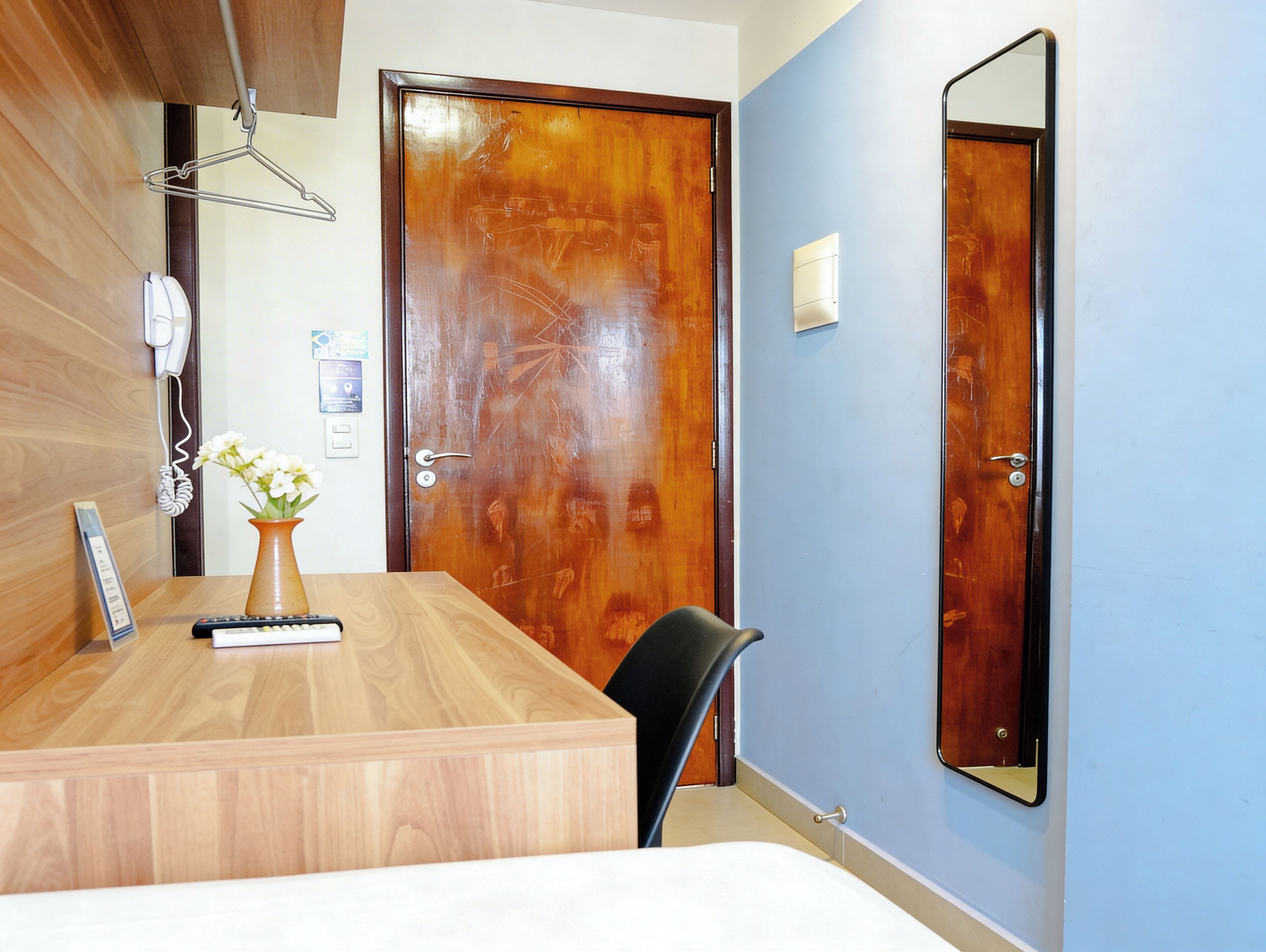 Ein Hotelzimmer mit einem Holztisch, einem Stuhl, einem Bett, einer Holztür, einem an der Wand befestigten Spiegel und einem an der Wand montierten Telefon.