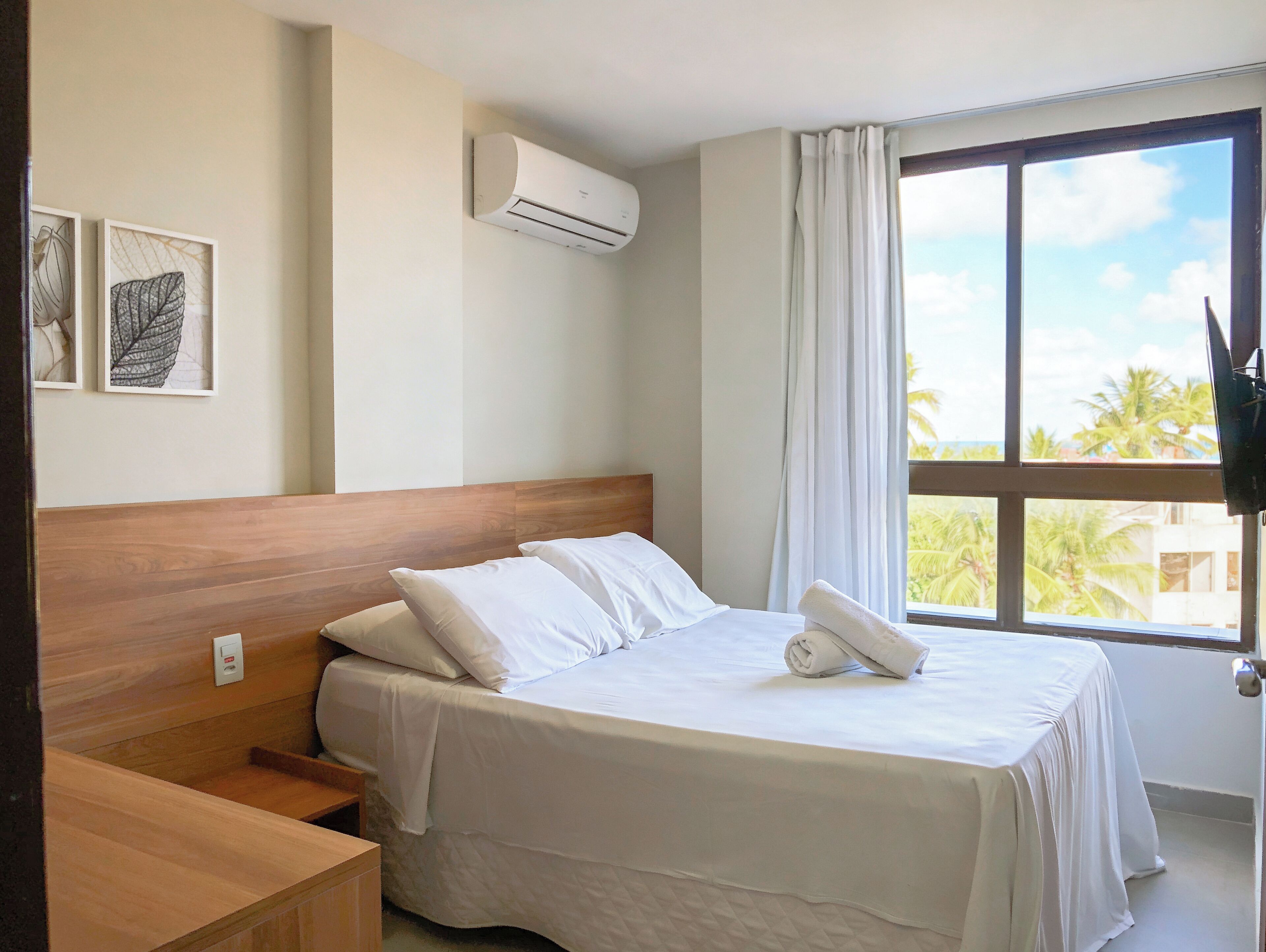 Una habitación de hotel con cama, mesita de noche de madera, aire acondicionado montado en la pared y ventana con vistas a palmeras.