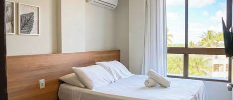 Una habitación de hotel con cama, mesita de noche de madera, aire acondicionado montado en la pared y ventana con vistas a palmeras.
