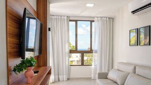 Un salón moderno con un televisor de pantalla plana montado en un panel de madera, un sofá beige, una mesita con un jarrón rosa y un ventanal con cortinas blancas.