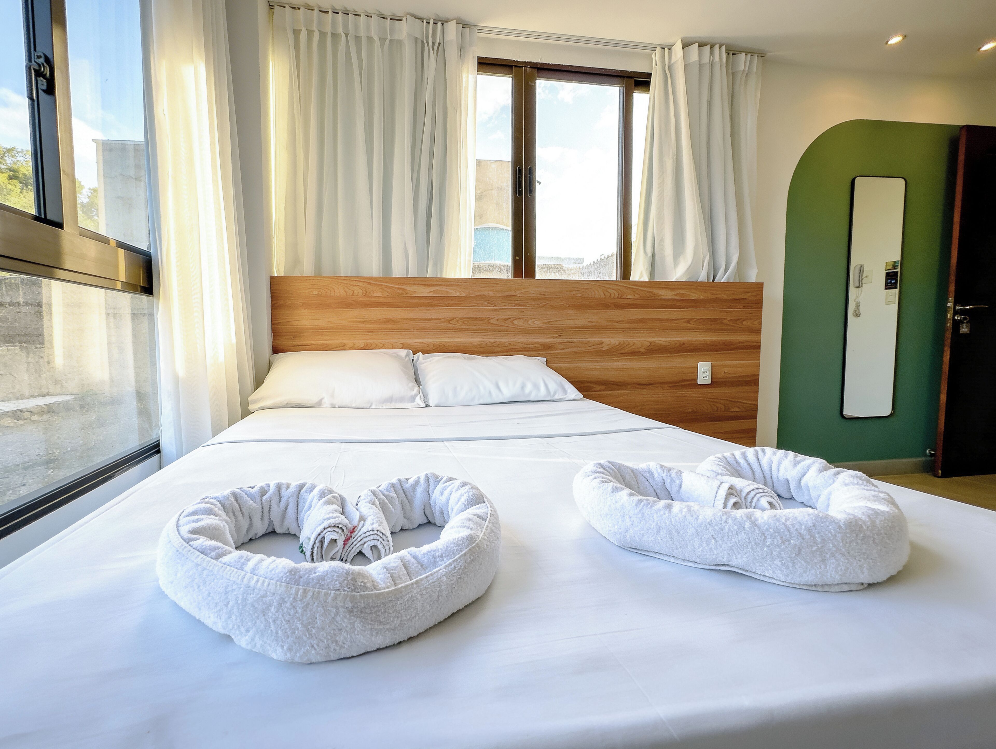 A hotel room with a large bed, two white towels in bed-shaped baskets, a wooden headboard, a green accent wall, and a window with curtains.
