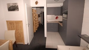 Een kleine keuken met grijze kastjes, een houten deur en een zithoek met een tafel en stoel.