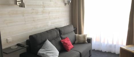 Ein modernes Hotelzimmer mit grauem Sofa, zwei Wandlampen, einem Holztisch und einem Fenster mit transparenten Vorhängen.