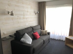 A modern hotel room with a grey sofa, two wall-mounted lamps, a wooden side table, and a window with sheer curtains.