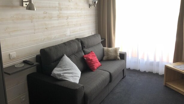 Een moderne hotelkamer met een grijze bank, twee wandlampen, een houten bijzettafeltje en een raam met dunne gordijnen.