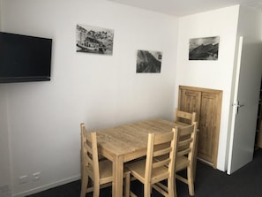 A room with a wooden table and chairs, a television mounted on the wall, and two black-and-white photographs.