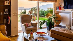 Una sala de estar con sofá, sillón y mesa de centro. Hay una chimenea, un televisor y una estantería. La habitación tiene un ventanal con vistas al exterior.