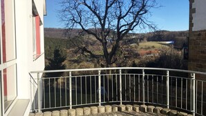 Ein Balkon mit Metallgeländer, ein Baum im Hintergrund und Blick auf eine hügelige Landschaft.