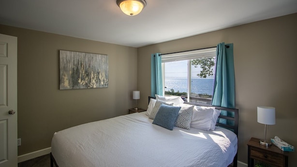 A bedroom with a large bed, a painting on the wall, a window with curtains, and a nightstand with a lamp. - Bayview House, Heirloom Vacations (Duluth)