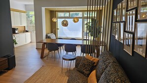 Una sala de estar moderna con sofá, mesa de comedor y sillas. Se aprecia una cocina al fondo, con armarios y una cafetera.