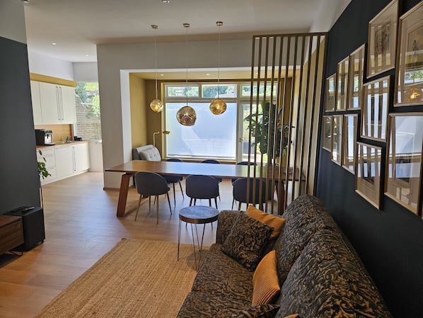 Un salon moderne avec un canapé, une table à manger et des chaises. On aperçoit une cuisine en arrière-plan, avec des placards et une machine à café.