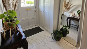 Un pasillo con una puerta blanca, un espejo que refleja la habitación, una araña y una planta en maceta.