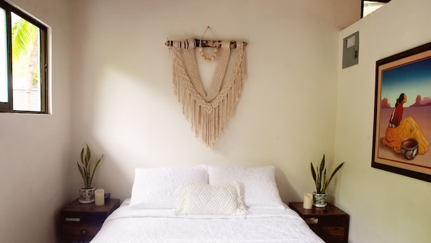 Un dormitorio con una cama, dos mesitas de noche, un colgador de macramé en la pared y dos plantas en macetas.