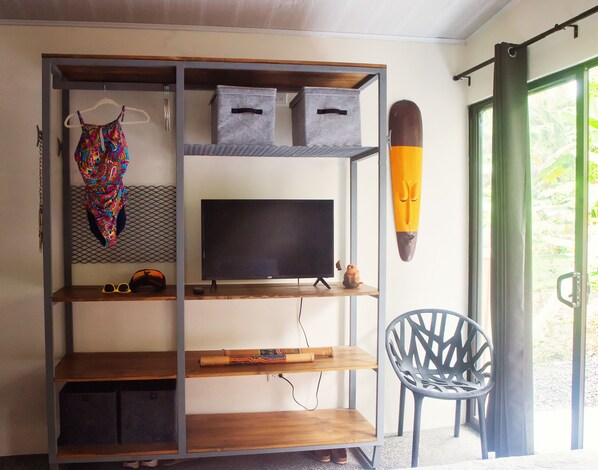 A room with a TV on a shelf, a chair, and a window with a view of greenery. - Beachside private home, walk to all. Fast wifi & pet friendly. (Guanacaste)