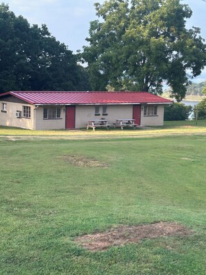 Edificio de una planta con techo rojo, dos ventanas y una puerta. Hay mesas de picnic frente al edificio.