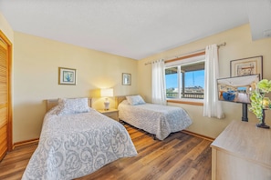 A hotel room with two beds, a wooden floor, a window with curtains, a television, and a small table with a plant. - Semi-Oceanfront Condo w/ Private Balcony, Resort Pool, Elevator, Beach Boardwalk (Rodanthe)